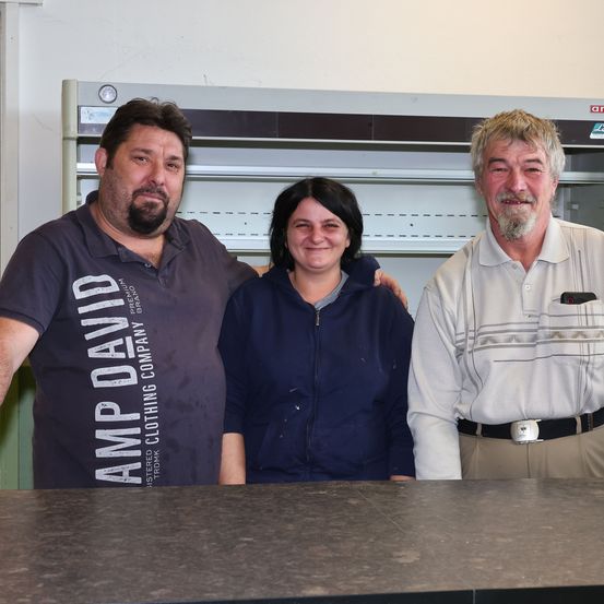 Drei Personen, zwei Männer und eine Frau, stehen zusammen in einem Raum und lächeln für ein Foto. Der Mann links trägt ein Shirt mit 'AMP DAVID' und 'CLOTHING COMPANY' darauf. Der Mann rechts trägt ein weißes Hemd und hat einen Gürtel. Die Frau ist in einer blauen Jacke. Hinter ihnen steht ein Regal mit Metallrahmen.