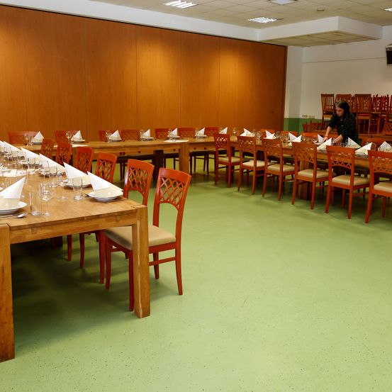 Ein Speisesaal mit mehreren Tischen, die für eine Mahlzeit gedeckt sind. Die Tische sind in Reihen angeordnet mit Stühlen. Jeder Tisch hat eine weiße Serviette, ein Glas und einen Teller. Eine Frau steht hinten, möglicherweise beim Einrichten des Tisches. Der Raum hat Holzwände, grünen Boden und Deckenleuchten.