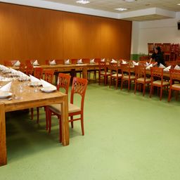 Ein Speisesaal mit mehreren Tischen, die für eine Mahlzeit gedeckt sind. Die Tische sind in Reihen angeordnet mit Stühlen. Jeder Tisch hat eine weiße Serviette, ein Glas und einen Teller. Eine Frau steht hinten, möglicherweise beim Einrichten des Tisches. Der Raum hat Holzwände, grünen Boden und Deckenleuchten.