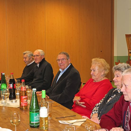 Eine Gruppe älterer Erwachsener sitzt um einen Tisch mit Weinläsern, Flaschen und Servietten. Eine Frau in einem roten Oberteil lächelt, während andere ernst aussehen. Hinter ihnen sind Holzwände und ein Holzstuhl zu sehen.
