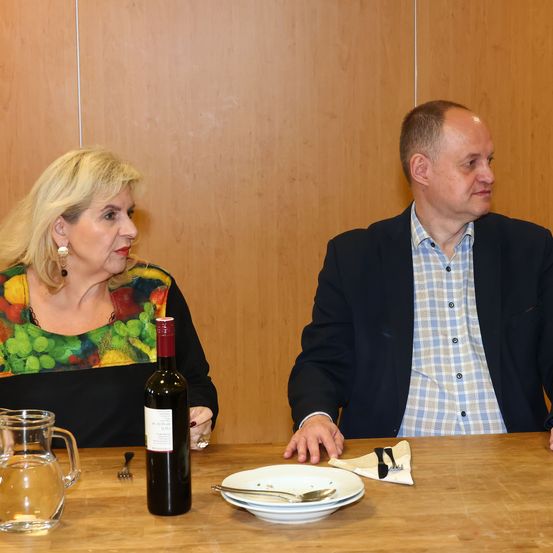Ein Mann und eine Frau sitzen an einem Tisch und sehen sich an. Die Frau hat eine Weinflasche und eine Karaffe. Der Mann hat ein Serviette und Besteck.