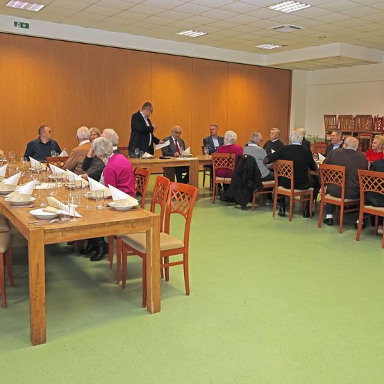 Eine Gruppe von Menschen versammelt sich in einem Speisesaal mit einer Holzwand und grünem Boden. Einige sitzen an Tischen, während andere in der Nähe eines Podiums stehen. Alle tragen formelle Kleidung.