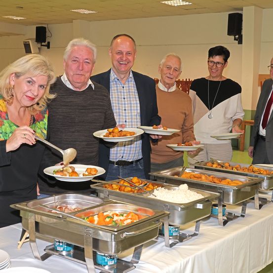 Eine Gruppe von Menschen steht hinter einem Buffettisch, lächelt und hält Teller mit Essen in der Hand. Sie befinden sich wahrscheinlich bei einer Gemeinschaftsveranstaltung. Der Tisch ist mit verschiedenen Gerichten gefüllt, und die Umgebung deutet auf eine gemeinschaftliche Zusammenkunft hin.
