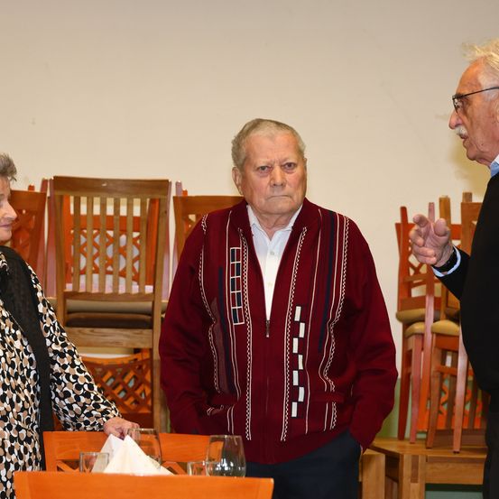 Drei ältere Erwachsene stehen in einem Raum mit Holzstühlen. Ein Mann mit Brille gestikuliert während er spricht. Ein anderer Mann trägt einen gemusterten Pullover. Eine Frau lächelt.
