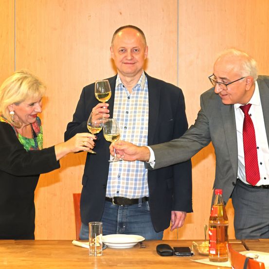 Drei Personen sind um einen Tisch versammelt und halten Weingläser in der Hand. Eine Frau links, ein Mann in der Mitte und ein älterer Mann rechts. Sie stoßen an. Der Tisch hat Teller, Gläser und eine Flasche. Der Hintergrund ist eine Holzwand.