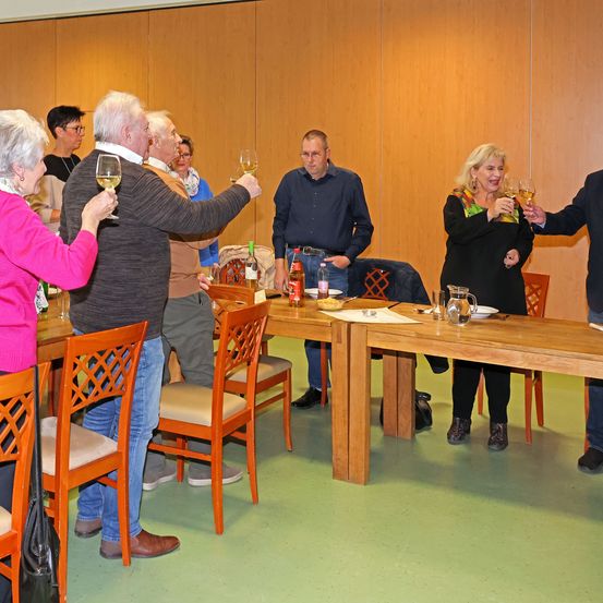 Eine Gruppe Erwachsener steht um einen Tisch herum, hält Weinglaser und scheint ein Ereignis zu feiern. Flaschen Wein und ein Krug stehen auf dem Tisch.
