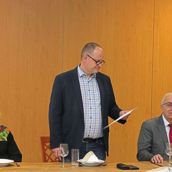 Ein Mann steht vor einer Holzwand und hält ein Papier in der Hand. Zwei Personen sitzen an einem Tisch, einer mit Serviette und Weinglas.