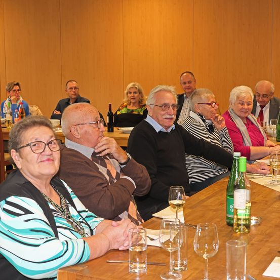 Eine Gruppe älterer Erwachsener sitzt in einem Besprechungsraum an einem Tisch. Sie führen Gespräche. Flaschen und Gläser stehen auf dem Tisch.