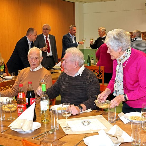 Mehrere ältere Männer und Frauen sitzen um einen Esstisch herum, wobei eine Frau Essen serviert. Dahinter halten andere Gäste Weingläser. Eine Holzwand und ein Holzboden sind sichtbar.