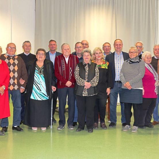 Eine Gruppe älterer Menschen, sowohl Männer als auch Frauen, stehen in einem Raum mit grünem Boden zusammen. Einige tragen Brillen und einer benutzt einen Stock. Sie sind in verschiedenen Winterkleidungen gekleidet und scheinen für ein Foto zu lächeln.