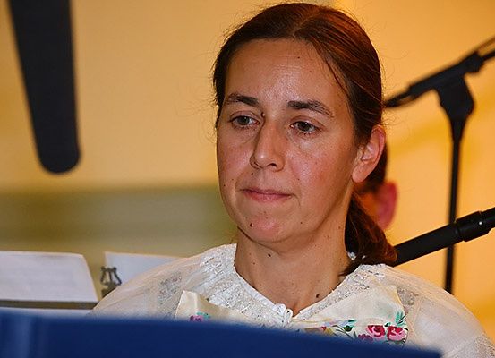 Eine Frau sitzt und liest von einem Notenständer. Hinter ihr ist ein Mikrofon auf einem Ständer zu sehen, und eine weitere Person steht verschwommen im Hintergrund.