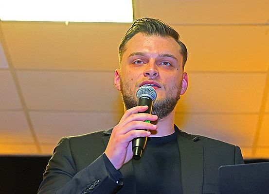 Ein Mann spricht bei einer Veranstaltung in ein Mikrofon, trägt einen schwarzen Anzug und hält das Mikrofon mit der linken Hand.