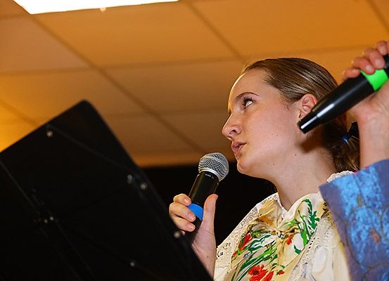 Eine Frau in einem Blumenhemd singt in ein Mikrofon und hält ein weiteres Mikrofon in der Hand. Sie hat einen ernsten Ausdruck.