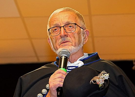 Ein älterer Mann mit Brille und weißem Bart spricht in ein Mikrofon. Er trägt einen schwarzen Umhang mit einem weißen Kragen und blauer Einfassung.