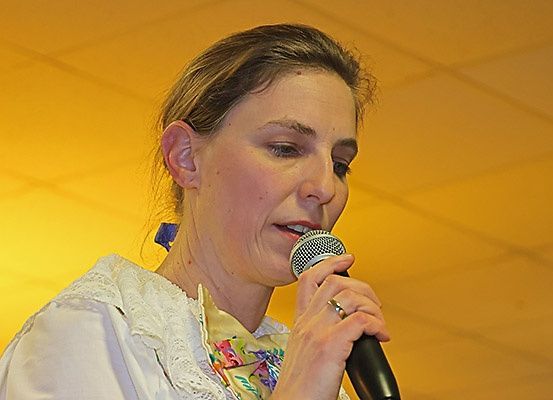 Eine Frau in einem weißen Blusenkleid mit Blumendesign hält ein Mikrofon und scheint bei einer Veranstaltung im Innenbereich zu singen.