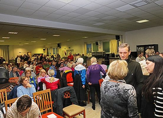 Eine große Gruppe von Menschen ist in einem Saal versammelt, einige sitzen und andere stehen. Ein Mann in einem schwarzen Anzug spricht zu der Menge. Der Saal hat Holzstühle, Jacken hängen an den Stühlen und Lautsprecher sind an der Wand montiert.
