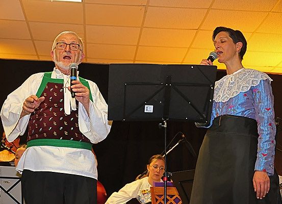 Ein älterer Mann und eine Frau singen auf der Bühne, mit einem Notenständer und einem Cellisten im Hintergrund.