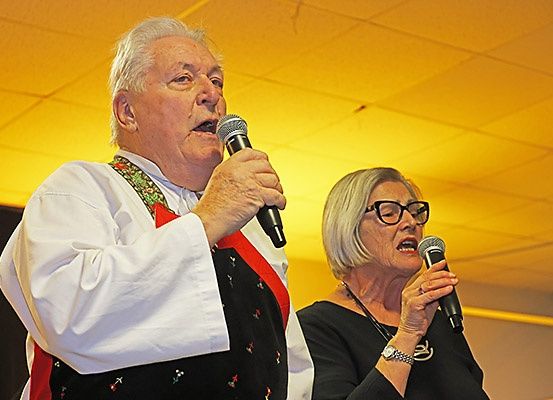 Ein älterer Mann und eine ältere Frau halten Mikrofone und singen in einem gelb beleuchteten Raum mit einer gelben Decke. Der Mann trägt ein weißes Hemd und eine dekorative Schärpe.