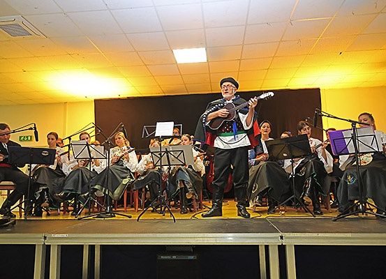 Ein Mann spielt Gitarre auf einer Bühne mit einer Band, beleuchtet von gelben Lichtern. Hinter ihm sitzen Musiker auf Stühlen und spielen verschiedene Instrumente. Ein schwarzer Hintergrund und ein Scheinwerfer sind vorhanden.