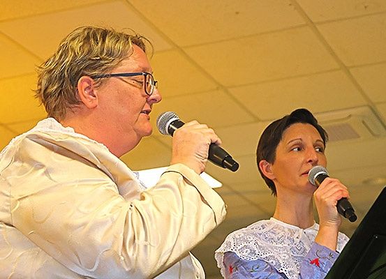 Zwei Frauen singen in Mikrofone. Eine Frau trägt eine Brille und einen beigefarbenen Mantel, die andere ist in einem weißen Spitzenkleid mit blauem Oberteil.