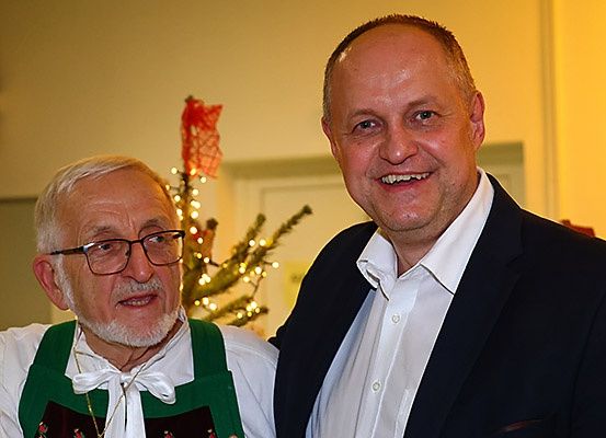Zwei Männer stehen zusammen mit einem lächelnden Gesicht. Der Mann auf der linken Seite trägt eine Brille und eine grüne Schürze. Hinter ihnen steht ein mit bunten Lichtern dekorierter Weihnachtsbaum.