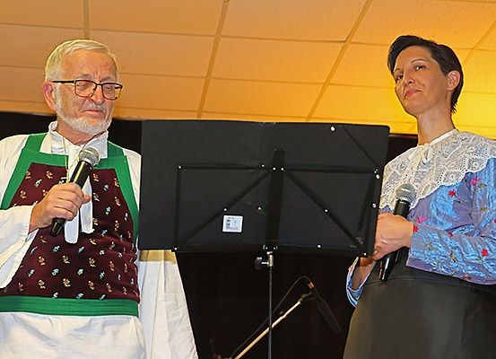 Ein älterer Mann und eine Frau stehen neben einem Notenständer mit Mikrofonen, wahrscheinlich bei einer Veranstaltung. Der Mann trägt eine Brille und eine traditionelle Weste.