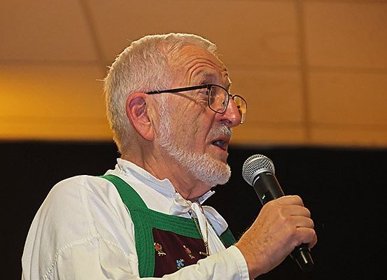 Ein älterer Mann mit weißen Haaren und Brille hält ein Mikrofon und spricht möglicherweise. Er trägt ein weißes Langarmhemd mit grüner Schürze.