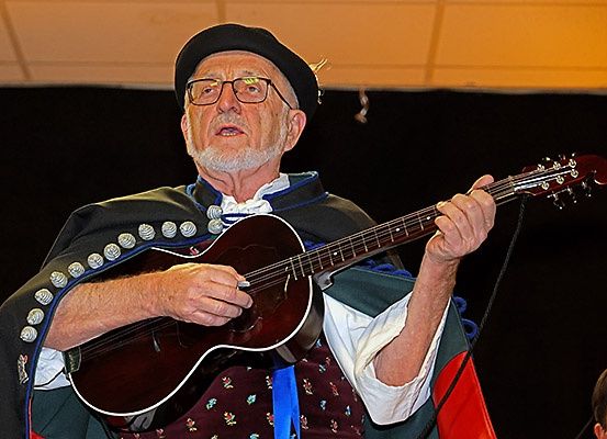Ein älterer Mann mit schwarzem Hut und Brille spielt Gitarre auf der Bühne, trägt einen Umhang mit Silberknöpfen und ein rotes Blumenwestchen. Er scheint in einem Konzert aufzutreten.
