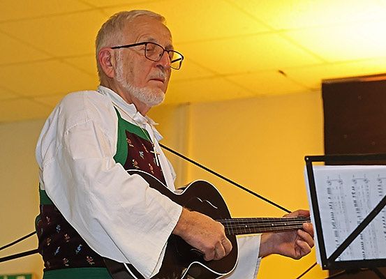 Ein älterer Mann mit Brille und traditioneller Kleidung spielt Gitarre und schaut auf Notenblätter.