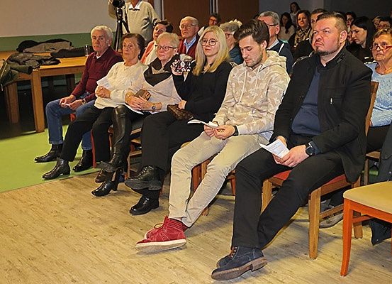Eine Gruppe von Menschen sitzt in einem Raum, einige mit Brillen. Einige schauen nach unten, möglicherweise lesen sie. Ein Mann steht in der Nähe eines Stativs. Kleidung und Taschen sind auf einem Tisch. Der Raum hat Holzböden.