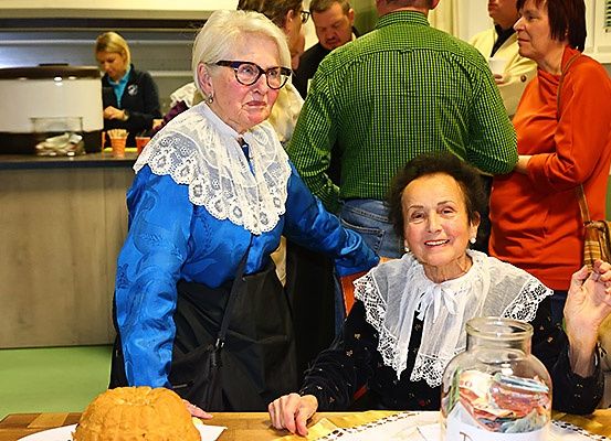 Zwei ältere Frauen, eine in Blau und eine in Schwarz, sitzen an einem Tisch mit Essen. Sie sind von anderen umgeben, möglicherweise bei einer Zusammenkunft oder Veranstaltung.