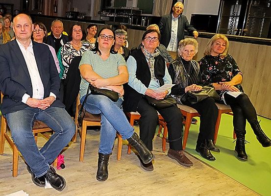 Eine Gruppe von Menschen sitzt in einem Raum mit Holzböden, einige tragen Brillen und Stiefel. Es gibt eine Theke mit einem Mann, der dahinter steht.
