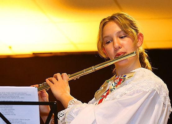 Ein Mädchen spielt eine Flöte und sieht nach unten. Sie hält die Flöte mit beiden Händen. Vor ihr liegt eine Musiknote. Sie trägt ein weißes Kleid mit einem roten und grünen Band. Das Licht scheint auf ihr Gesicht.