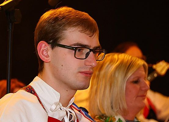 Ein Mann mit Brille schaut etwas vor sich an. Er trägt ein weißes Hemd mit roten und blauen Bändern. Hinter ihm steht eine Frau mit einer grünen Blumenkette.