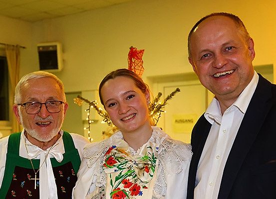 Drei Personen posieren für ein Foto; ein älterer Mann, eine Frau in traditioneller Kleidung und ein Mann in formeller Kleidung. Hinter ihnen ist ein festlicher Hintergrund mit Dekorationen.