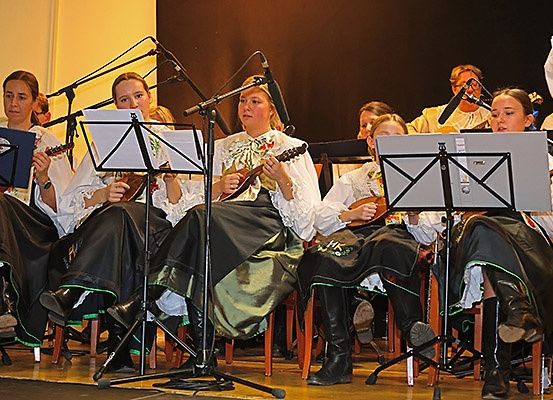 Eine Gruppe von Frauen spielt auf der Bühne Saiteninstrumente, trägt traditionelle Kostüme und sitzt auf Stühlen.