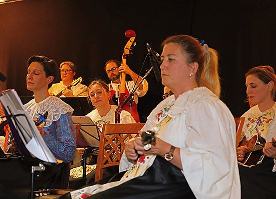 Eine Gruppe von Musikern, gekleidet in traditioneller Kleidung, tritt auf der Bühne auf. Sie spielen verschiedene Instrumente, darunter Gitarren und einen Cello. Eine Frau vorne trägt ein weißes Kleid und hält eine Gitarre.