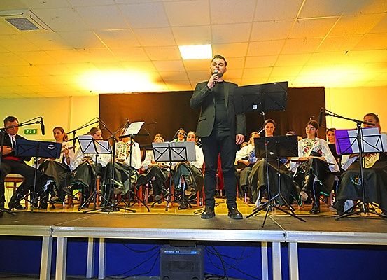 Ein Mann singt auf der Bühne mit einem Mikrofon, begleitet von einer Gruppe Musiker, die verschiedene Instrumente spielen. Das Publikum sitzt auf Stühlen hinter ihnen, unter einer Lampe.