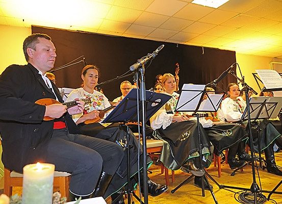 Ein Mann spielt eine Gitarre vor einer sitzenden Gruppe von Musikern mit Notenblättern und Mikrofonen.