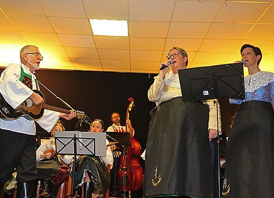 Ein älterer Mann spielt Gitarre auf der Bühne, während zwei Frauen singen. Ein Cellist und ein Violinist sitzen im Hintergrund.