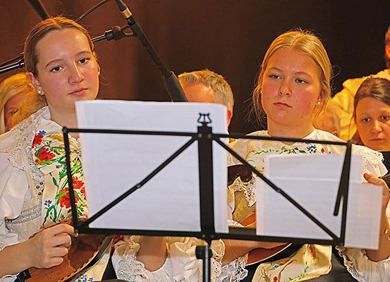 Zwei Frauen spielen Gitarren bei einem Konzert, gekleidet in traditionelle Kostüme. Ein Notenständer hält Notenblätter vor ihnen. Im Hintergrund sind andere Künstler und ein verschwommenes Publikum zu sehen.