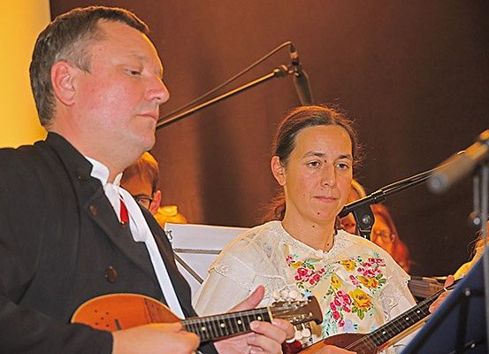 Ein Mann und eine Frau spielen Gitarren bei einem Konzert. Sie stehen vor einem Mikrofon. Die Frau trägt ein Blumenkleid. Hinter ihnen sitzen andere Leute auf Stühlen, und es gibt eine gelbe Wand.