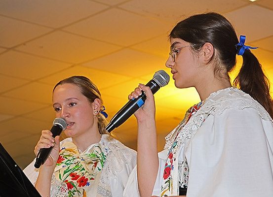 Zwei Mädchen in traditioneller Kleidung singen in Mikrofone, eines schaut in die Kamera. Sie tragen eine Brille und einen Pferdeschwanz mit blauen Schleifen. Dahinter steht ein Notenständer.
