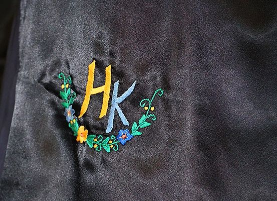 Ein Nahaufnahme eines schwarzen Stoffes mit den gestickten Buchstaben HK, umgeben von bunten Blumen und Blättern.