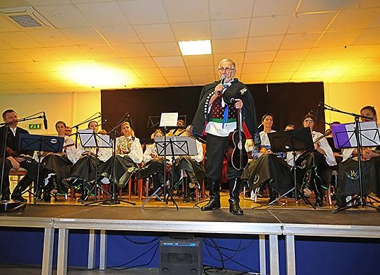 Ein älterer Mann steht mit einem Mikrofon auf der Bühne und spricht vor einem Publikum. Hinter ihm spielt eine vielfältige Gruppe von Musikern verschiedene Instrumente. Sie sitzen in Reihen, dem Publikum zugewandt, mit Notenständern vor ihnen.