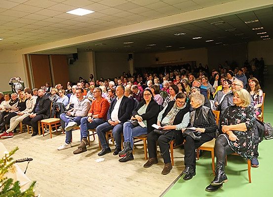 Ein großes Publikum sitzt in Reihen in einem Raum mit Holzboden, grünem Teppich und einem Weihnachtsbaum auf der linken Seite. Sie schauen sich eine Veranstaltung an, und einige halten Papiere in den Händen.