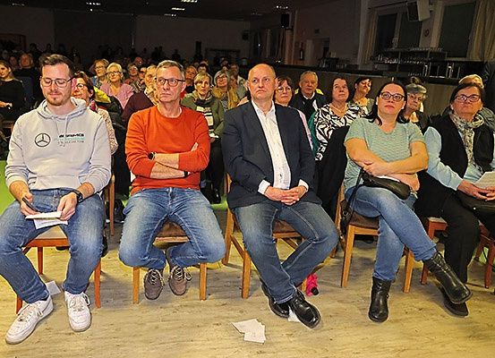 Mehrere Menschen sitzen in einem Konferenzsaal, einige mit Brillen und Turnschuhen, einer hält ein Papier. Zwei Männer vorn tragen rote und graue Hemden, einer mit verschränkten Armen. Eine Frau vorne trägt Stiefel. Der Raum ist holzfarben und mit Menschen gefüllt.