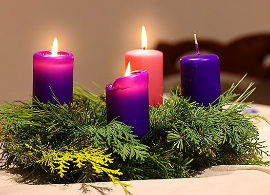 Vier brennende Kerzen, lila und rosa, sind in einem Kranz aus grünen Blättern auf einem Tisch platziert. Ein gelber Zweig ist im Vordergrund.