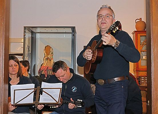 Eine Gruppe von Menschen spielt Gitarren. Ein Mann steht und spielt. Eine Frau sitzt und liest eine Musikpartitur. Dahinter steht eine Vitrine mit einer Puppe darin.