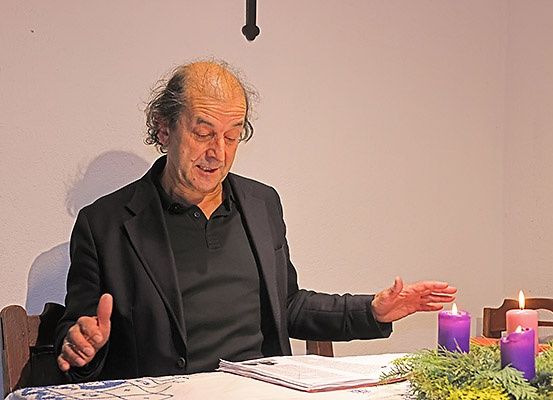 Ein älterer Mann sitzt an einem Tisch und gibt eine Daumen hoch. Kerzen und ein Kranz schmücken den Tisch, mit Stühlen drumherum. Er trägt einen schwarzen Anzug und hat ein Buch vor sich offen.